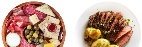 A wooden platter with assorted meats, cheeses, crackers, olives, and a small jar of jam sits next to a white plate of sliced steak and potatoes garnished with green onions.